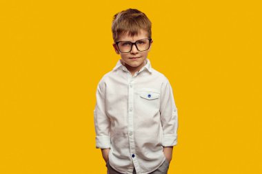 Beyaz gömlek ve gözlük takan, elini cebinde tutan ve sarı arka planda kameraya kızgın bakan ciddi bir çocuk.