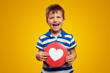 Mavi çizgili polo giyen gülümseyen çocuk, sosyal ağlar için kalp ikonu gösterirken aynı zamanda sarı arka planda duruyor.