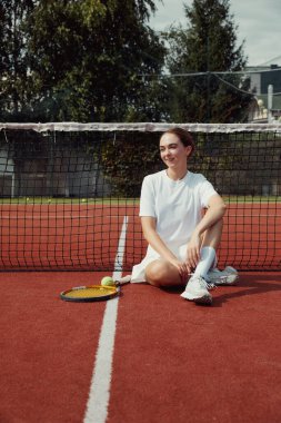 Beyaz tişörtlü ve elbiseli, tenis kortunda raketle tenis sahasında oturan çekici bir sporcunun dikey fotoğrafı..