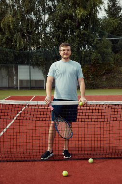 Tenis raketi tutan, tenis sahasında poz veren genç bir adamın dikey fotoğrafı. Hafta sonları ve pazar günleri dinlenmek için. Arkadaşlarla aktif spor oyunu.