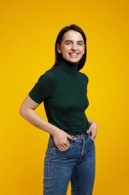 Heyecanlı esmer kızın dikey portresi, yeşil tişört giyiyor, gülümsüyor ve ceplerinde el ele tutuşuyor, sarı arka planda izole edilmiş.