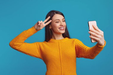 Mavi arka planda izole edilmiş, elinde akıllı bir telefonla barış jesti yapan havalı, neşeli bir kızın yatay fotoğrafı.