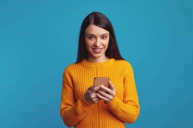 Cep telefonu kullanan, banka hesabını kontrol eden ya da sosyal ağlarda mesaj gönderen, turuncu kıyafetler giyen, mavi arka planda izole edilmiş mutlu genç kız.