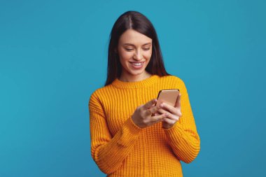 Akıllı telefon kullanan, banka hesabını kontrol eden ya da sosyal ağlarda mesaj gönderen, turuncu kıyafetler giyen, mavi arka planda izole edilmiş gülümseyen bir kadın.