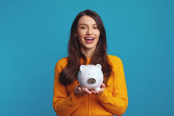 stock image Cheerful young girl in casual orange sweater smiling and holding white piggy bank with lots of money isolated over blue background