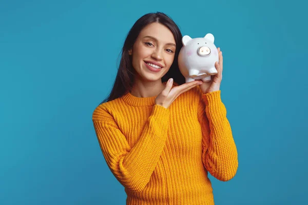 stock image Attractive young female in knitted orange sweater holding white piggy bank with lots of money near face isolated over blue background