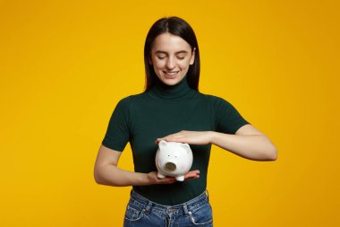 Sarı arka planda kumbarası olan, yeşil tişört giyen, para kutusunu ararken gülümseyen çekici bir kadın.