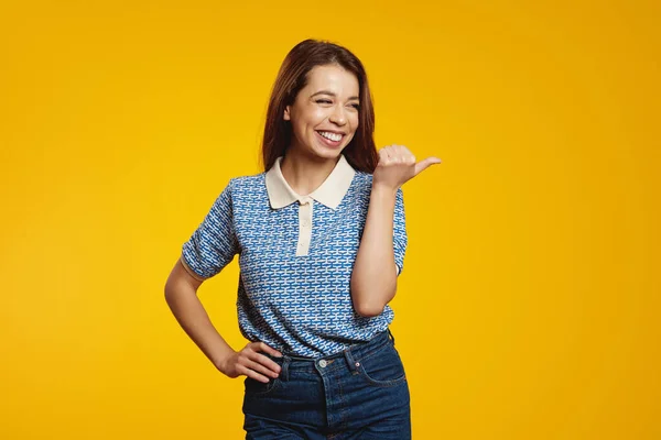 stock image Cheerful smiling woman pointing fingers at copy space and looking away while smiling, standing against yellow background. Advertising concept.