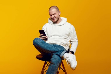 Sandalyede oturan heyecanlı adam, modern akıllı telefonda mobil uygulama kullanıyor, arkadaşlarıyla sohbet ediyor ya da komik videolar izliyor, sarı arka plan