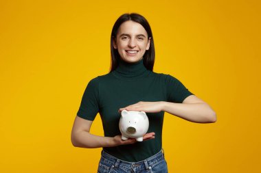 Sarı arka planda kumbarası olan, yeşil tişört giyen, kameraya bakarken gülümseyen çekici bir kadın. Para kutusu