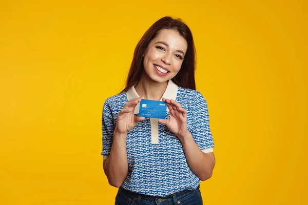 Stock image Smiling pretty brunette girl holding credit card while smiling and looking at camera over yellow background, wearing blue shirt