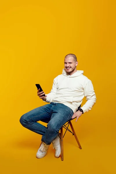 Jovem Sorrindo Usando Smartphone Enquanto Sentado Cadeira Isolado Fundo Amarelo — Fotografia de Stock