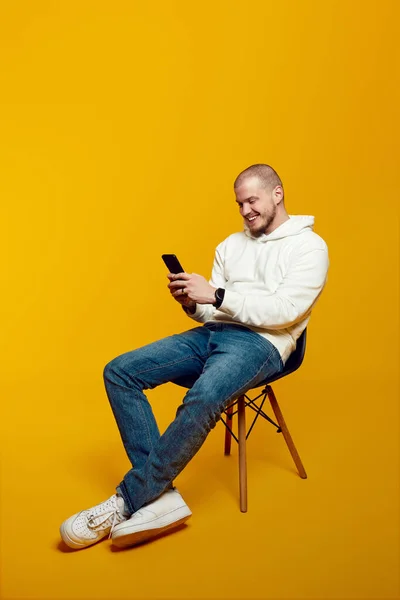노란색 배경에 고립된 의자에 스마트폰을 사용하여 잘생긴 남성을 것입니다 신나는 — 스톡 사진