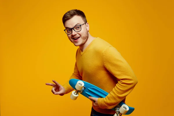 Szczęśliwy Młody Człowiek Żółtym Swetrze Okularach Trzymając Niebieski Longboard Pokazując — Zdjęcie stockowe