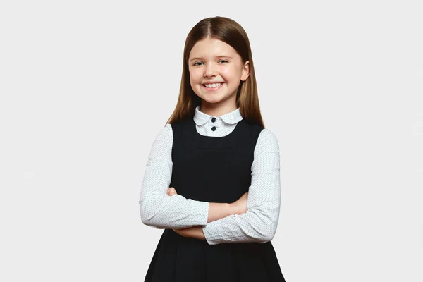 Sarı saçlı, güzel ilkokul öğrencisi kız ellerini çapraz yapıp beyaz arka planda dişlek tebessümle kameraya bakıyor.