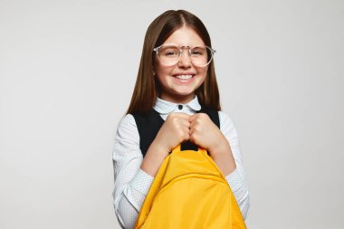 Okul çalışmalarından önce kameraya gülümseyen ve sarı sırt çantasıyla beyaz arka planda izole edilmiş neşeli okul çocuklarının yakın portresini çek.