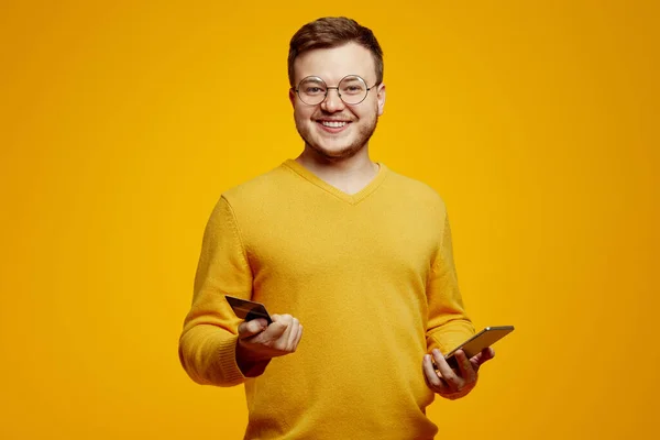 턱수염 덥수룩 청년은 주황색 스웨터와 안경을 있으며 스마트폰 카드를 있으며 — 스톡 사진