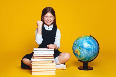Neşeli liseli kız, sarı arka planda izole bir şekilde, zaferi kutlamak için yumruğunu kaldırırken bir sürü kitabın ve toprak küresinin yanında oturuyor.