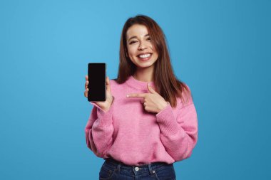 Mavi arka planda soyutlanmış, boş ekrana bakarken gülümseyen ve akıllı telefon tutan pembe kazaklı mutlu genç bir kadın.