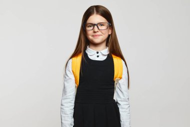 Güzel görünümlü inek öğrenci gözlüklü, okul üniformalı ve sarı sırt çantalı beyaz arka planda kameraya bakıyor. Okul konseptine dönelim.