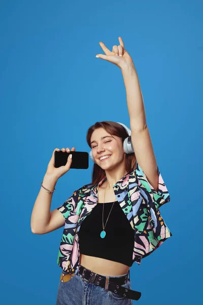 照片上是一个快乐的小女孩 一边拿着耳机一边听音乐 一边拿着智能手机 展示着摇滚的标志 蓝色背景与之隔离 情感和生活方式概念 — 图库照片