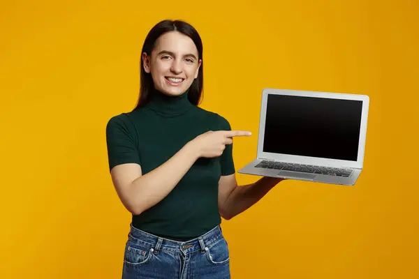 stock image Trendy hipster girl wearing green t-shirt pointing at new laptop in hands, isolated over yellow background.