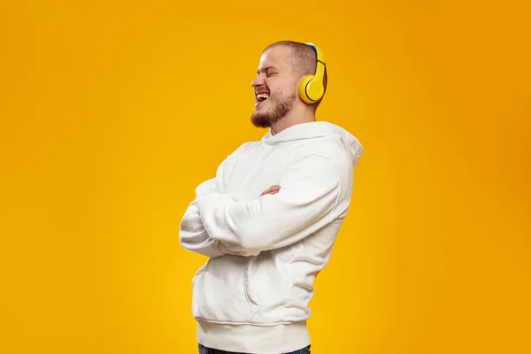 stock image Smiling attractive man in white hoodie, listening to music in yellow headphone with closed eyes while holding hands crossed against orange background