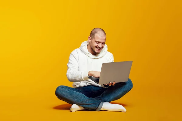 Genç adam yerde oturuyor ve modern not defteri kullanıyor, dizüstü bilgisayarda yazıyor ve gülümsüyor, sarı arka planda izole edilmiş, mesaj için boş bir alan var.