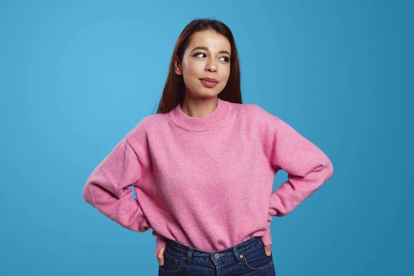 ピンクのセーターを着た陽気な若いヒスパニック系の女性は 青い背景に目を背けながら 腰に手を置いて立っています 人々感情の概念 — ストック写真