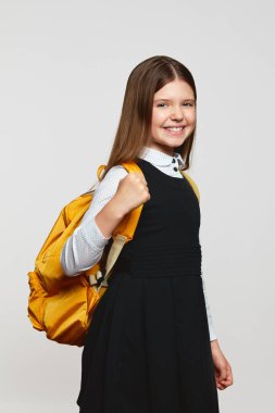 Sarı sırt çantalı ve kollu bir kızın dikey fotoğrafı beyazlara karşı başarıyı kutlarken gülümsüyor. Okul konseptine dönelim.