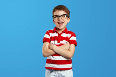 Heyecanlı inek çocuk kırmızı çizgili gömlek ve gözlük takıyor kolları mavi arka planda ve kameraya bakıyor.
