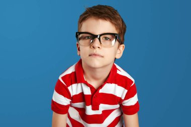 Kırmızı çizgili polo gömlek ve mavi arka planda izole edilmiş gözlük takan, bunalmış inek bir gencin yakın plan portresi. Negatif insan duyguları, yüz ifadeleri, duygular
