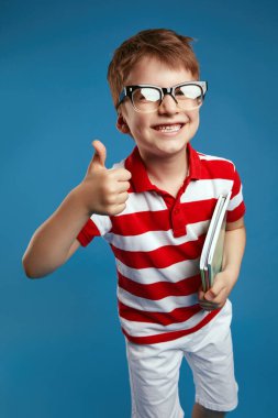 Retro gözlüklü, kırmızı çizgili tişörtlü, elinde ders kitabı olan inek bir çocuğun portresini kapat. Baş parmak hareketini göster. Mavi arka planda tek başına dur.