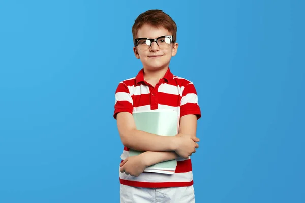 快乐的小男孩 戴着书呆子眼镜 拿着记事本 站在蓝色背景下 穿着红色条纹马球衫 对着相机微笑 — 图库照片