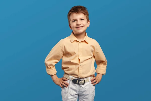 ベージュシャツを着たハッピーな小さな少年は 青い背景に対して手を挙げながら カメラのために微笑みます 子供のスタジオの肖像画 子供時代のライフスタイルコンセプト — ストック写真
