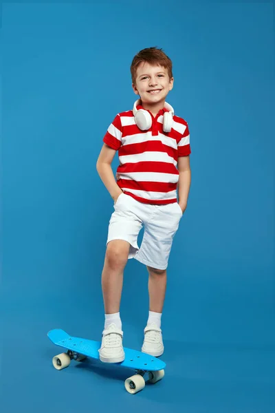 男孩脖子上穿着红色条纹马球衫 头戴耳机 在滑板上抱着腿 在蓝色背景下对着相机微笑的全身上下照片 — 图库照片