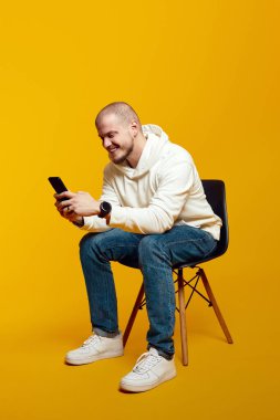 Sakallı genç erkek, sarı arka planda izole sandalyeye otururken akıllı telefon kullanıyor. Heyecanlı adam internette sohbet ediyor, sosyal medyada geziniyor.