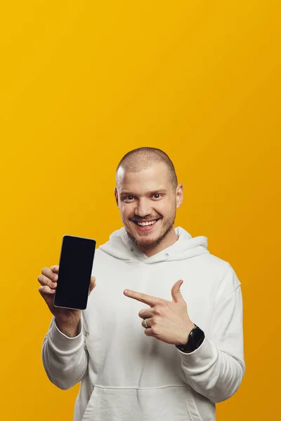 黄色の背景に隔離された白いフーディーを着て笑っている間白いスクリーンの携帯電話を指す幸せな若者の縦の写真 — ストック写真
