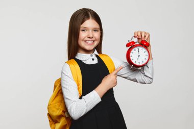 Okul üniformalı neşeli inek kız ve sarı sırt çantalı beyaz arka planda kırmızı alarmlı saati işaret ediyor. Okula geç kalma..