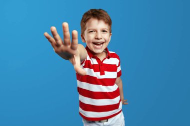 Çizgili kırmızı polo giyen mutlu küçük çocuk, mutlulukla gülüyor ve kameraya uzanıyor. Stüdyo görüntüsü, blu arkaplan üzerinde izole
