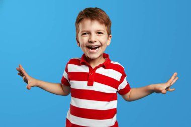 Kırmızı çizgili polo tişörtlü mutlu çocuk mavi arka planda kollarını kaldırarak kutluyor. Çocuk stüdyo resmi.
