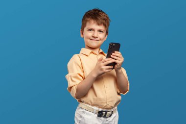 Bej tişört giyen, elinde akıllı telefon tutan ve gülümseyen, cep telefonu kullanan neşeli, masum bir çocuk. Stüdyo çekimi mavi arkaplanda izole edildi