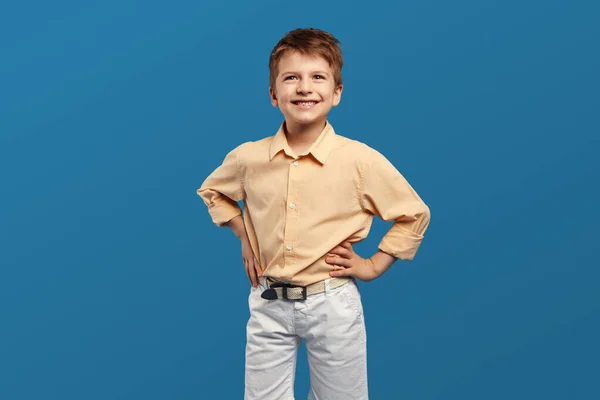 エキサイティングなハンサムな男の子はベージュシャツを着て 青い背景に対して手を挙げながらカメラのために微笑みます 子供のスタジオの肖像画 子供時代のライフスタイルコンセプト — ストック写真
