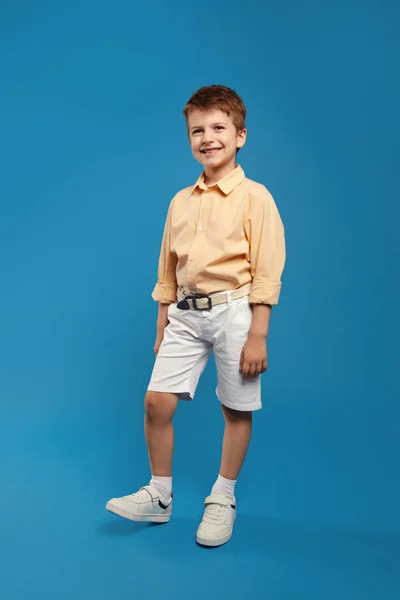 照片上 一个英俊的小男孩穿着米黄色的衬衫 一边对着相机微笑 一边手挽手靠着蓝色的背景图片 童年生活方式概念 — 图库照片