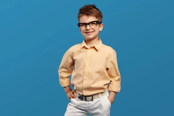 スマートな小さな男の子は 青い背景に対して腰を保ちながら カメラのために笑顔で神経質な眼鏡とベージュのシャツを着ています 子供スタジオの肖像画 — ストック写真