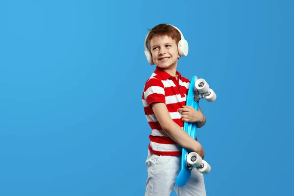 学龄前小男孩身穿红色条纹衬衫 手持滑板 通过耳机听音乐 同时寻找空余的文字空间 用蓝色背景隔开 — 图库照片