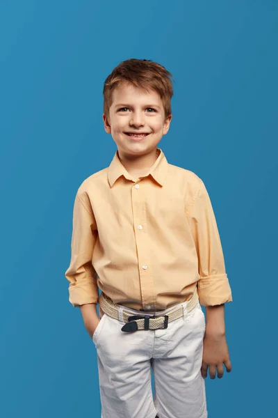 一个英俊可爱的小男孩穿着时髦的米黄色衬衫 一边对着相机微笑 一边手插在蓝色背景的口袋里的垂直照片 童年生活方式概念 — 图库照片