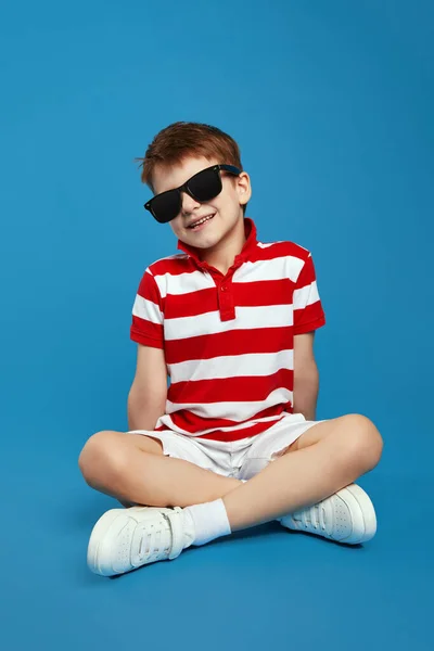 캐주얼 선글라스에 귀여운 소년의 사진은 파란색 배경에 앉아서 카메라를보고 로열티 프리 스톡 사진
