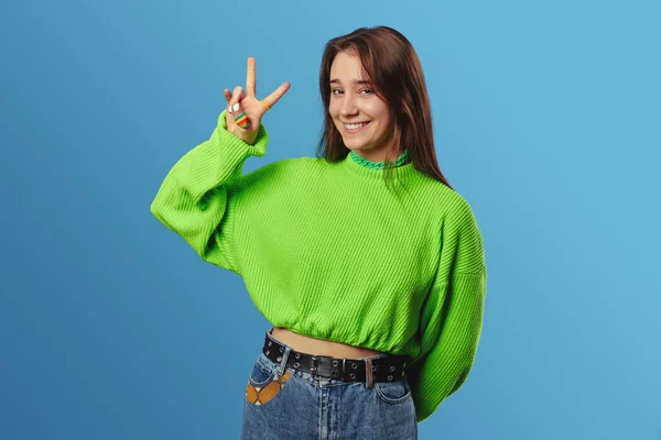 身穿绿色毛衣的年轻漂亮女士 面带微笑 在蓝色背景的相机前摆出V字牌 — 图库照片
