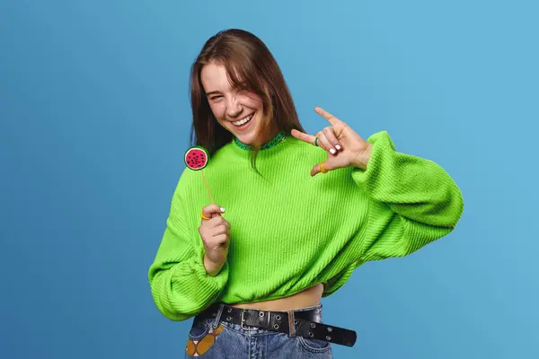 穿着绿色毛衣和牛仔裤的快乐的年轻女性 拿着糖果 带着蓝色背景 微笑着 摆出摇滚的姿势 — 图库照片